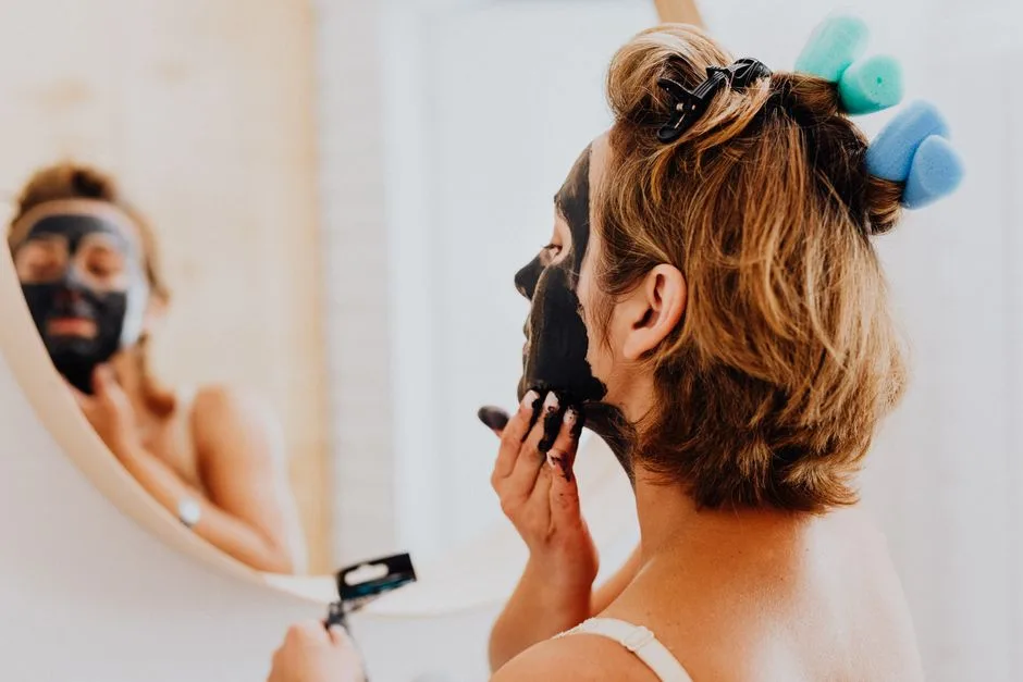 A woman is applying a black facial mask in front of a mirror, focusing on skincare and beauty routines.