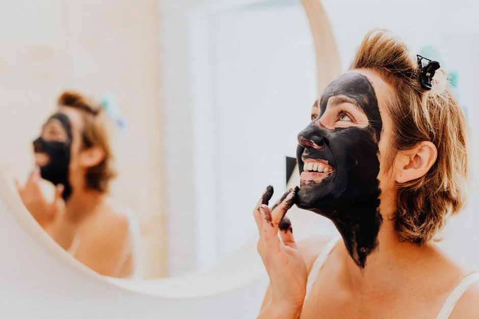 Happy woman applying black charcoal face mask in front of a mirror.